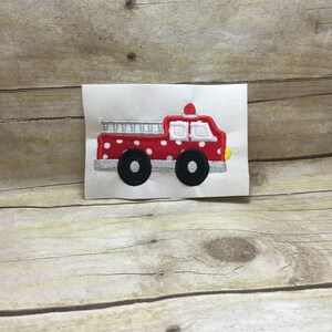 May include: Embroidered patch of a red fire truck with white polka dots, black wheels, and a silver ladder. The fire truck has a red light on top and yellow headlights. The patch is on a white background.