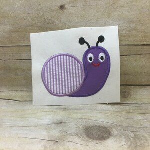 Puede incluir: Aplique de caracol bordado de color morado con una concha rayada y una cara sonriente.