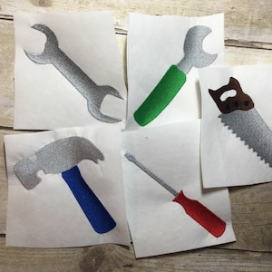 May include: Five iron-on patches featuring tools: a wrench with a green handle, a wrench with a silver handle, a saw with a brown handle, a hammer with a blue handle, and a screwdriver with a red handle.