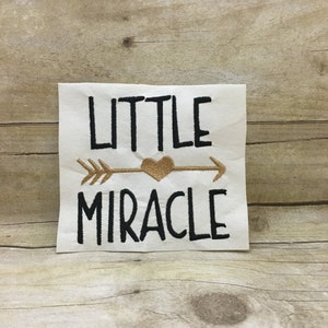 May include: White fabric with black embroidery that reads "LITTLE" with a gold arrow and heart, and "MIRACLE" underneath.