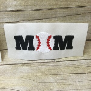 May include: White fabric with the word "MOM" embroidered in black with a red and white baseball in the centre.