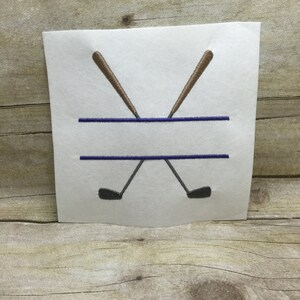 May include: A white embroidery design with two crossed golf clubs in brown and a blue line for text.