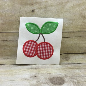 Könnte beinhalten: Rote und weiße Gingham-Kirschen mit grünen Blättern und weißen Punkten. Die Kirschen sind auf einem weißen Stoff gestickt.