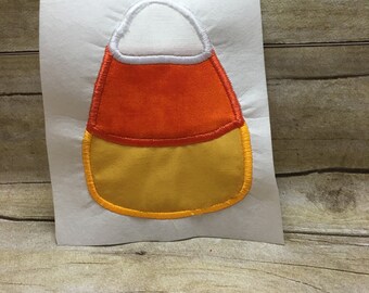 Op de afbeelding: Een vilt applique in de vorm van een snoepmais met een witte bovenkant, een oranje midden en een gele onderkant. De applique heeft een gele omtrek.