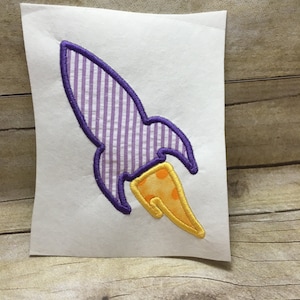 Könnte beinhalten: Lila und gelbe gestickte Raketenapplikation mit weißen Streifen am Körper und gelben Punkten auf der Flamme.