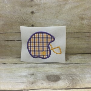 May include: Purple and gold plaid football helmet applique with a yellow circle in the center.