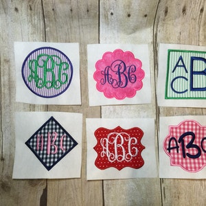 Peut inclure: Six carrés de tissu blanc avec des monogrammes brodés de couleurs et de motifs différents. Les monogrammes sont tous "ABC" dans différentes polices et styles. Les carrés sont sur un fond en bois.