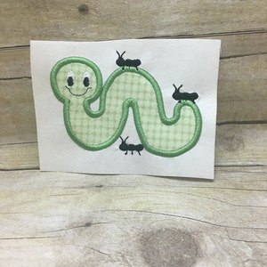 May include: Green embroidered caterpillar with a smiling face and three black ants crawling on it.