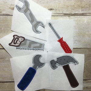 May include: Five iron-on patches featuring tools: a wrench, a saw, a screwdriver, a wrench with a blue handle, and a hammer. The patches are on white fabric.