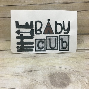 May include: Embroidered design on white fabric with the text "Baby Cub" in black and gray letters. A small brown tent is between the words "Baby" and "Cub".