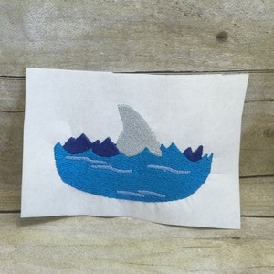 May include: Embroidered design of a shark fin sticking out of the water. The water is blue with white waves and the fin is grey.