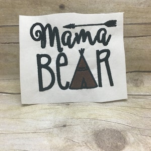 Pode incluir: Tecido branco com bordado preto que lê "Mama Bear" com uma seta acima da palavra "Mama" e uma tenda marrom abaixo da palavra "Bear".