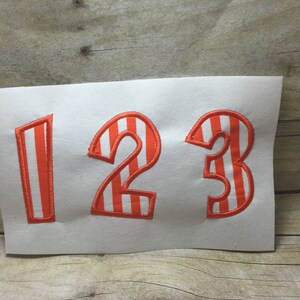 Puede incluir: Tres números de fieltro de rayas naranja y blancas, 1, 2 y 3, sobre un fondo blanco.