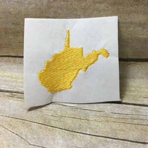 May include: A gold embroidered outline of the state of West Virginia on a white background.