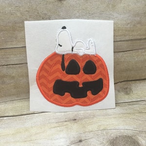 May include: An orange and black embroidered pumpkin with a white embroidered Snoopy sleeping on top.