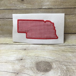 Puede incluir: Tela de vichy roja y blanca en forma del estado de Nebraska. La tela está cosida con un contorno rojo.