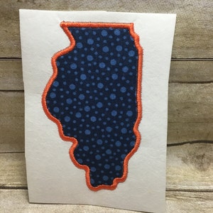 Op de afbeelding: Een blauwe en oranje stoffen applicatie van de staat Illinois. De applicatie is op een witte achtergrond.