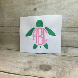 May include: A white fabric patch with a green embroidered sea turtle with a pink monogram "ABC" in the center.