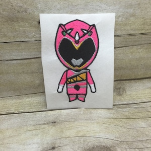 May include: Pink Power Ranger character embroidered on a white background. The character is wearing a pink helmet with a gold and silver design. The character is also wearing a pink suit with a gold and silver belt.