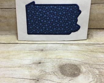 Könnte beinhalten: Ein blauer und weißer Stoff-Patch in Form des Bundesstaates Pennsylvania. Der Patch hat ein Punkte-Muster.