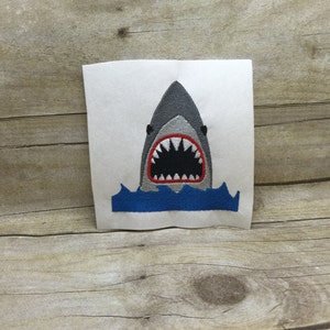 Pode incluir: Etiqueta bordada de um tubarão cinzento com a boca aberta e dentes vermelhos, sobre uma onda azul.