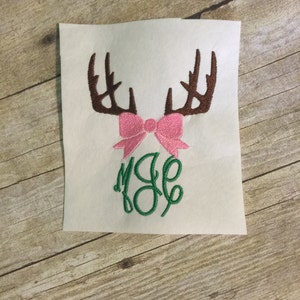 May include: Embroidered design featuring a pair of brown antlers with a pink bow and the monogram "MJE" in green.