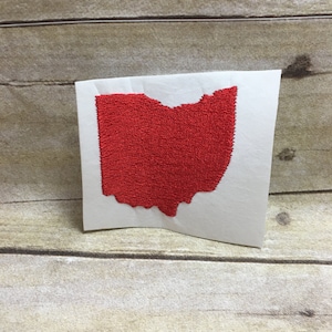 Puede incluir: Un parche bordado rojo con la forma del estado de Ohio, sobre un soporte blanco. El parche está sobre un fondo de madera clara. El bordado rojo tiene una apariencia texturizada.