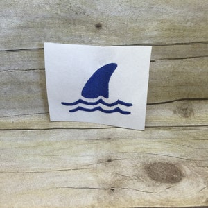 May include: Blue shark fin embroidered patch with white waves. The patch is on a white background.