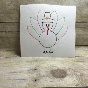 Könnte beinhalten: Ein gestickter weißer Truthahn mit einem roten Kehllappen, grünen und orangefarbenen Federn und einem braunen Pilgerhut. Der Truthahn ist auf einem weißen Hintergrund gestickt.