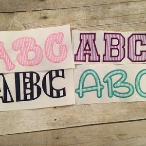 May include: Four different styles of embroidered letters, "ABC", in pink and white gingham, purple and white gingham, navy and white stripes, and teal and white gingham.