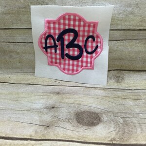 May include: A pink and white gingham fabric monogram with the letters "ABC" in navy blue.