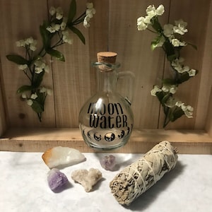 May include: A clear glass bottle with a cork stopper and a handle, labeled "Moon Water". The bottle is surrounded by white sage smudge sticks, crystals, and white flowers.