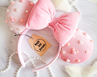 Peut inclure: Oreilles de souris Minnie en velours rose avec un grand nœud rose et des embellissements en perles. Les oreilles ont un serre-tête avec le texte "Magic And Dreams" sur une étiquette.