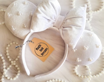 May include: White velvet Minnie Mouse ears with a large white bow and pearl embellishments. The ears have a headband with a brown tag that says "Magic And Dreams".