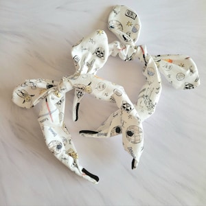 May include: Two white fabric headbands with black elastic. The fabric has a white background with black and white illustrations of Star Wars characters and spaceships. The headbands are tied in a bow.