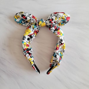 May include: A white headband with a colorful print of Mickey Mouse and friends. The headband is tied in a bow at the top.