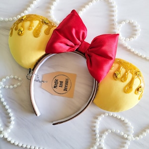 May include: Yellow Minnie Mouse ears with a red bow and dripping honey detail. The ears are decorated with small, gold bees. The headband is brown with a white inner band. The text "Magic And Dreams" is printed on a brown tag.