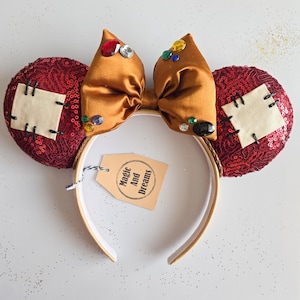 May include: A decorative headband with sequined red ears, a gold satin bow, and colorful jewel accents. The ears feature a stitched patch design. A tag with the text "Magic And Dreams" is attached to the headband.