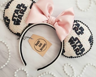 May include: A pair of white and black mouse ears with a pink bow. The ears are decorated with a white background and black text that says "Star Wars".