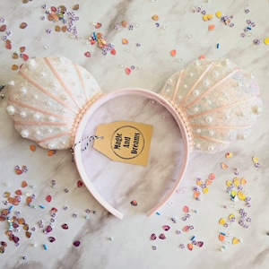 May include: A pair of pink and white mouse ears with a pearl and sequin design. The ears are attached to a pink headband. The headband has a brown tag with the text "Magic And Dreams".