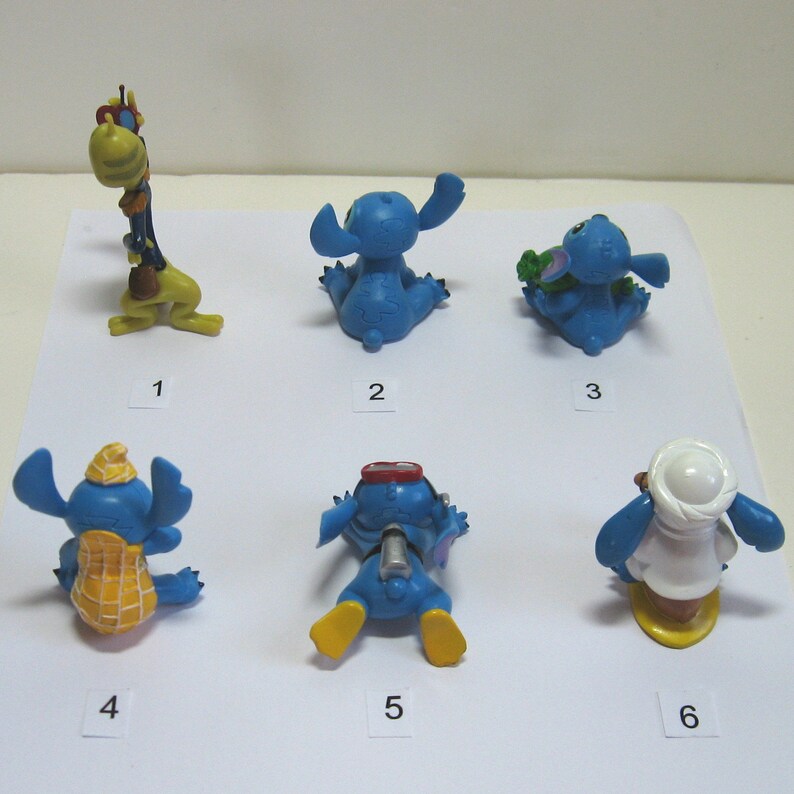 K&ouml;nnte beinhalten: Eine Sammlung von sechs kleinen, blauen Stitch-Figuren in verschiedenen Posen. Eine ist eine Giraffe, und die anderen tragen verschiedene Outfits oder halten Gegenst&auml;nde. Die Figuren sind mit 1 bis 6 nummeriert.
