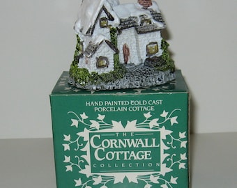 Może przedstawiać: Ręcznie malowana figurka domku z zimowym krajobrazem wykonana z zimnego odlewu porcelany. Figurka stoi na zielonym pudełku z napisem "The Cornwall Cottage Collection" i "Winter Cottage".