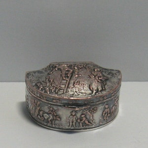 May include: A silver and copper trinket box with a raised design of people and a ladder. The box is round with a scalloped top.