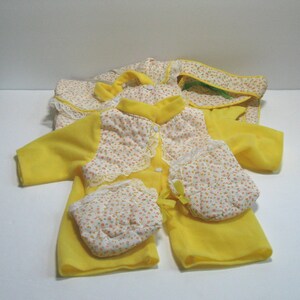 May include: A yellow baby outfit with a floral print and lace trim. The outfit includes a jacket, pants, and a pair of mittens.