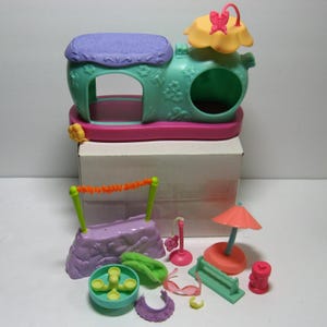 May include: A colorful toy playset featuring a teal and purple house-shaped structure with a flower and butterfly accent. Additional accessories include a swing set, bench, umbrella, and other miniature items. The set is designed for imaginative play.