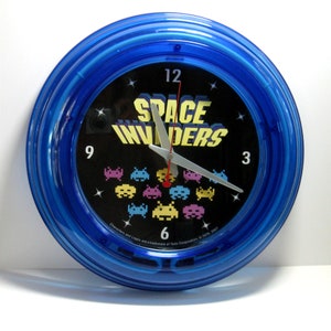 May include: A blue neon clock with a black face featuring the words "Space Invaders" in yellow and white letters. The clock face also has a graphic of the classic video game Space Invaders with colorful alien spaceships. The clock hands are silver.