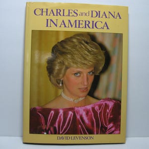 May include: A book titled "CHARLES and DIANA IN AMERICA" with a portrait of Princess Diana on the cover. The cover is yellow with purple text and a photo of Princess Diana wearing a pearl necklace and earrings.
