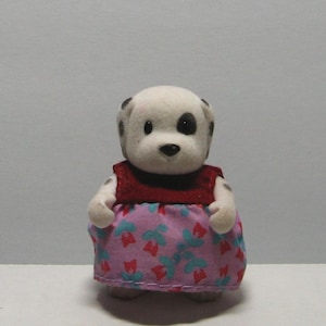 May include: A small, white Dalmatian figurine with black spots on its head and body. It wears a red top and a pink dress with a floral pattern. The toy is designed with a soft, flocked texture.