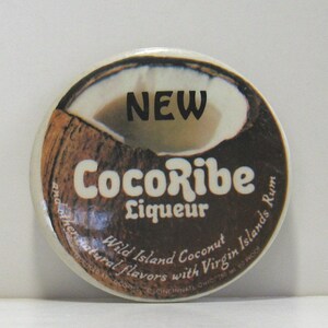 May include: A round, brown and white button with the text "NEW CocoRibe Liqueur Wild Island Coconut and other natural flavors with Virgin Islands Rum" printed on it.