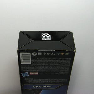 May include: A black cardboard box with the Marvel logo and the words "IRON MAN" in blue. The box has recycling symbols and text in multiple languages. The box is on a white background.